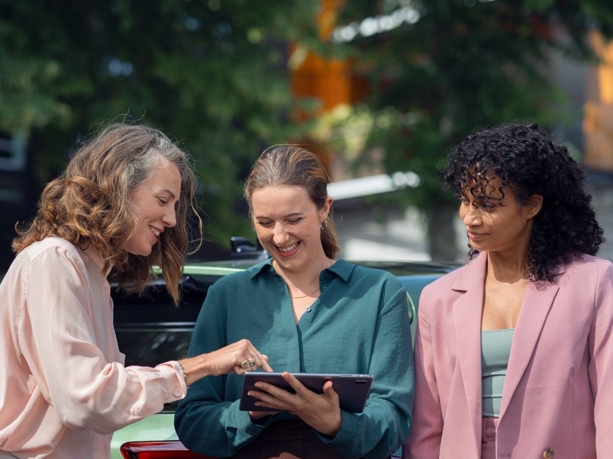 Three women discuss in front of a green car, holding a tablet.