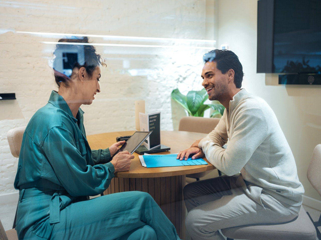 Two people sitting at a round wooden table in an office, discussing documents and a tablet. 