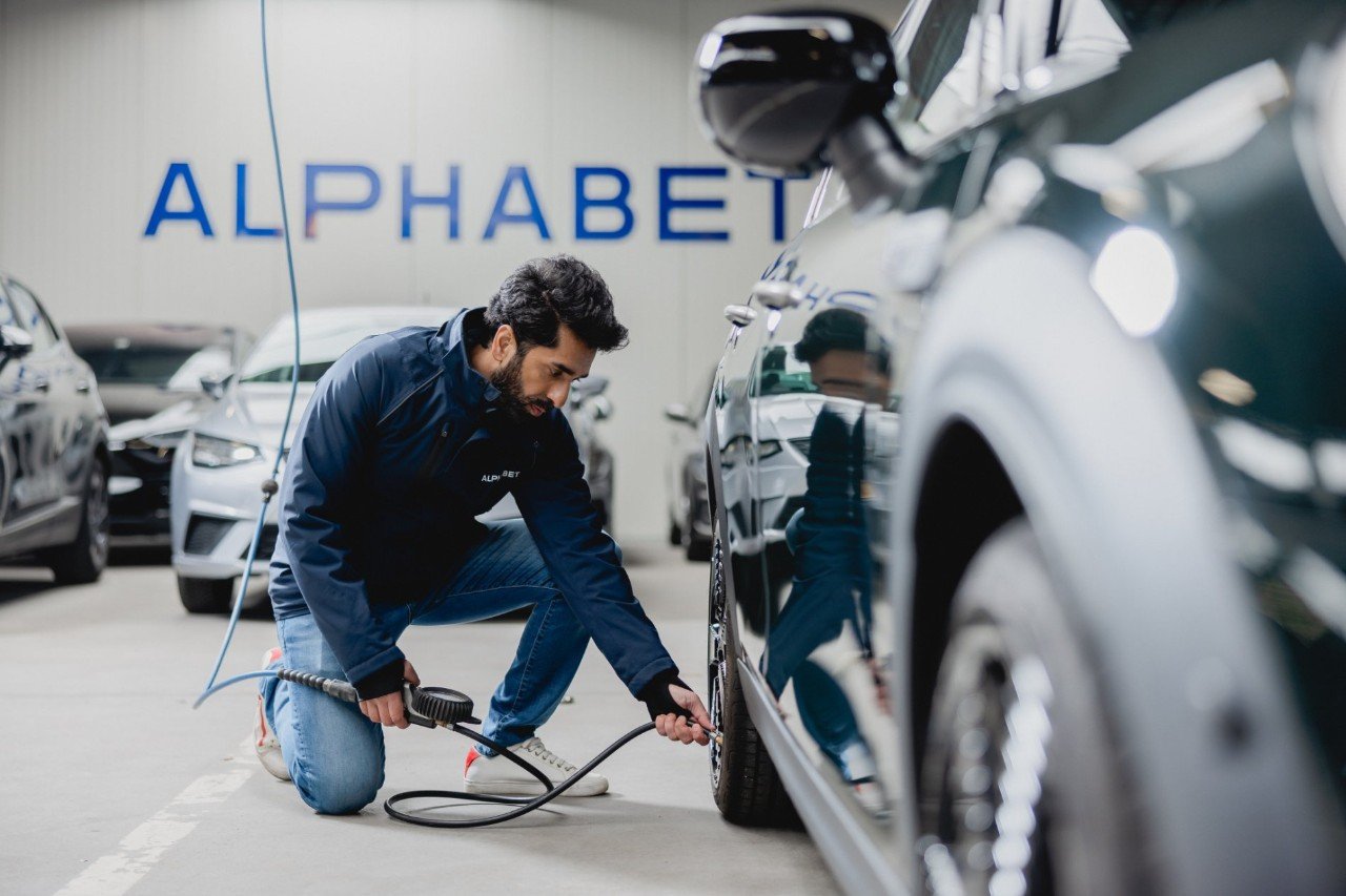 An Alphabet employee checks and inflates a car tire in a service area with parked vehicles.